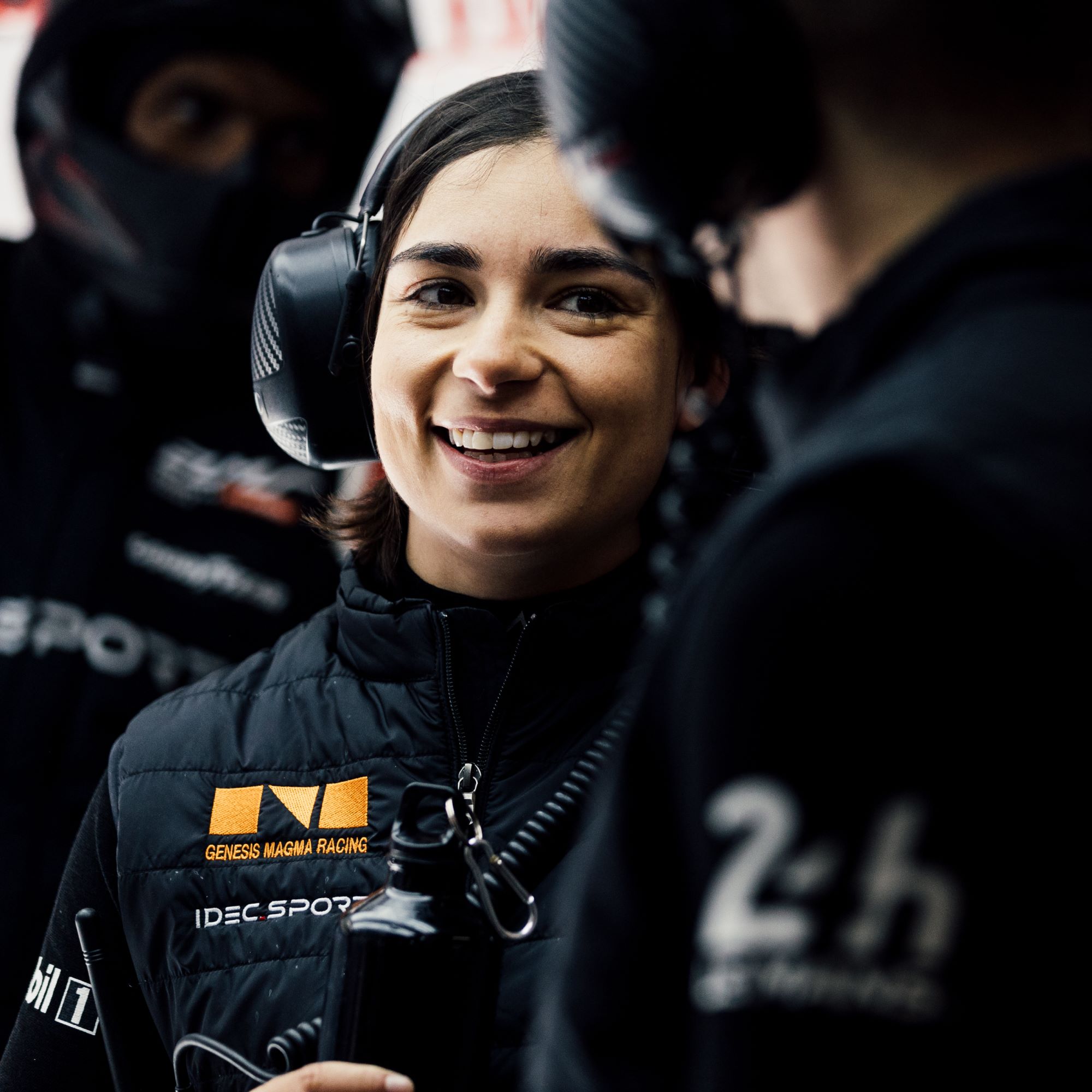 Driver Jamie Chadwick smiles wearing headphones and a black racing vest. Driver Jamie Chadwick smiles wearing headphones and a black racing vest.